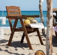 An outdoor chair with armrests,  on the beach,  shown together with different sorts of melons in a bowl.